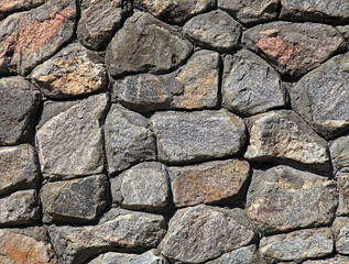 Granite wall, background. Surface of stone structure built of granite rocks (bricks). The wall texture of volcanic grey color. Fence with large gray stones on cement base in the rays of the sun