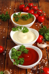 There are three bowls of mayonnaise, ketchup and mustard on a wooden table.