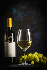 A dark, elegant still life of white wine in a glass with wine bottles and fresh grapes in the background. Perfect for wine-themed compositions