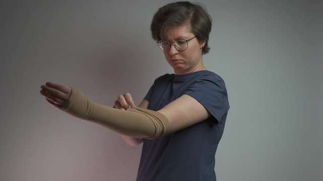 Prophylactics of thrombosis. Woman removes compression sleeve from left hand on gray background. Effective anti-thrombosis therapy involves compression sleeves to improve vein health and blood flow. 