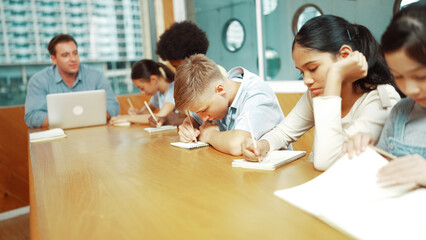 Young smart teenager writing and talking about topic at classroom. Diverse highschool student taking a note in other notebook while sitting with multicultural friends. Creative education. Edification.