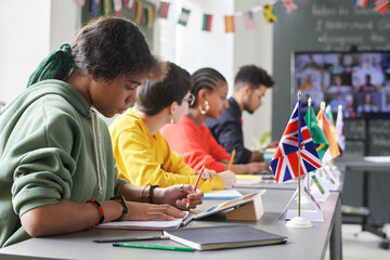 Side view at diverse group of students sitting at table with country flags during international conference in school