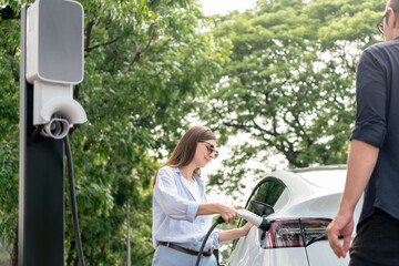 Naklejka premium Lovely young couple wearing sun glasses recharging battery for electric car during road trip travel EV car in natural forest or national park. Eco friendly travel during vacation and holiday. Exalt