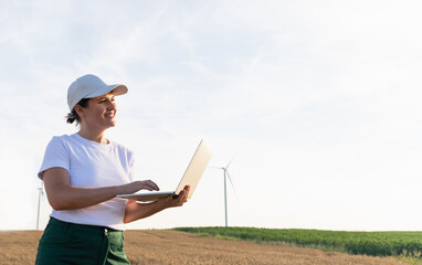 Woman farmer wearing white cap and t-shirt with laptop. Wind turbines in the background.