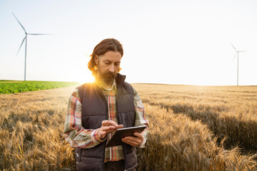 Farmer with digital tablet works on an agricultural field at sunset. Wind turbines in the background