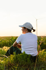 Woman wearing white cap siWoman wearing white cap sits on the field at sunset. Wind turbines in the background..ts next to solar panel at sunset. Wind turbines in the background.