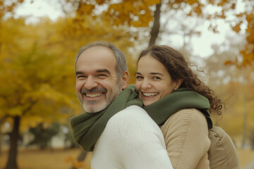 Fototapeta premium A smiling middle-aged man carrying a woman on his back, wearing a white sweater and green scarf; a park in the background; autumn