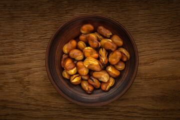 roasted broad beans with salt in a clay bowl in the center of the frame on a textured wooden table. top view