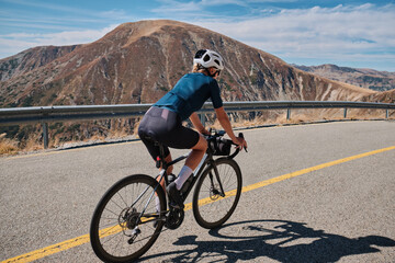 Naklejka premium A woman rides her bike on a winding road, surrounded by stunning mountain views. Female cyclist is wearing a blue jersey and black shorts, with a helmet on her head.