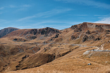 Stunning panoramic view of a winding road through a mountain pass. Rugged peaks, dry grass, and a clear blue sky create a picturesque scene. Parâng Mountains, Romania. Transalpina road.