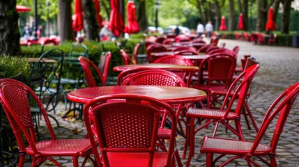 An outdoor café scene featuring bright red metal tables and chairs, set against a green backdrop of trees and bushes. The bold color of the furniture contrasts with the natural greenery,