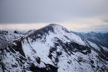 Stunning aerial view of snow-capped mountain peaks with a hint of blue sky peeking through the clouds. Perfect for nature, landscape, and winter themes. Fagaras mountains, Romania - Powered by Adobe