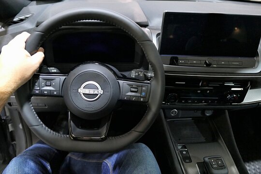 Driving wheel and detail of dashboard of modern japanese compact crossover SUV car Nissan Qashqai, third generation with 2024 facelift, hand of driver and legs visible.