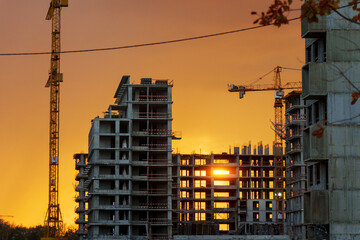 Golden sunset behind a bustling construction site with cranes and buildings silhouetted against the...