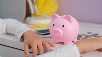 Close-up of young girl sits at her table, carefully dropping coins into her piggy bank, focusing on budgeting and learning the value of saving money