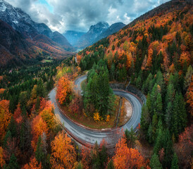 Aerial view of winding mountain road in forest at sunset in autumn. Alps, Italy. Curved alpine road, snowy rocks, trees, red leaves, dramatic cloudy sky with clouds. Colorful. Top view of road in fall