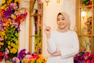 Young woman hijab wearing long white dress muslim cheerful to camera hands folded pointing right top among flower decoration inside luxury restaurant. for fashion, beauty, lifestyle content