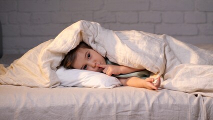 Little girl hiding under blanket making silence gesture