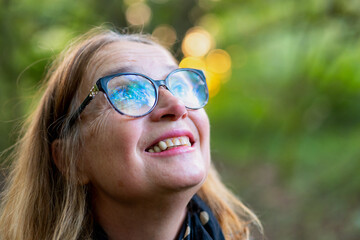 Woman smiling and looking upwards, wearing glasses. The background is out of focus, with a natural,...