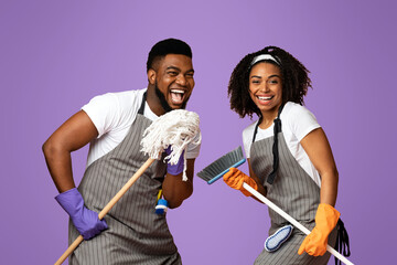 Positive african couple fooling together while making spring cleaning, singing into mop and having fun over pink background with copy space