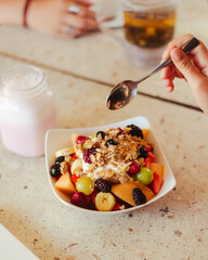 Mano sosteniendo una cuchara, debajo un bowl con frutos rojos y cereales de un desayuno saludable