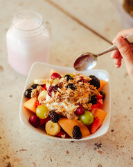 Mano sosteniendo una cuchara, debajo un bowl con frutos rojos y cereales de un desayuno saludable