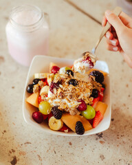 Mano sosteniendo una cuchara, debajo un bowl con frutos rojos y cereales de un desayuno saludable