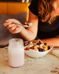 Mano sosteniendo una cuchara con cereales y yogurt, debajo un bowl con cereales, frutas y un vaso de smoothie refrescante. 