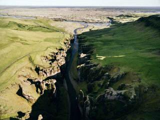 Drohenperspektive von einer bewachsener grüner Schlucht mit einem Fluss und einer offenen Ebene im Hintergrund, Island, Fjaðrárgljúfur