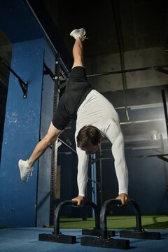 Vertical shot of muscular man in handstand position with one leg on side mastering gymnastic technique on floor planche bars in gym, copy space