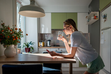 Morning procrastination of woman awaked looking at phone screen, drinking water, reading news, scrolling social media web standing in kitchen. Relaxed female spends time alone before breakfast at home