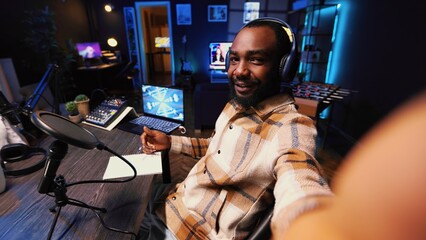 Journalist records interview with smartphone selfie camera in studio, reviewing notes to ask quality questions to guest. POV shot of BIPOC man using phone to film vlog, talking on mic with woman