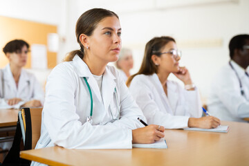 Interested young female doctor trainee in white coat listening to lecture on general medical...