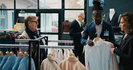 Clothing store employee makes fashion recommendations for client, helping senior woman to choose elegant shirt and suit jacket. Team of assistants suggesting trendy clothing items. Camera B.