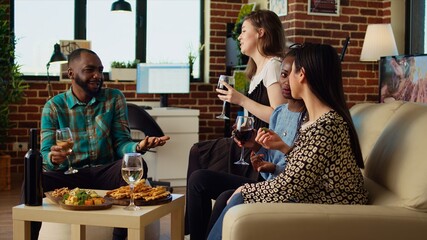 Culturally diverse friends talking, drinking alcoholic beverages and eating cheese, bread sticks from charcuterie board at home. Multicultural colleagues enjoying social life during weekend