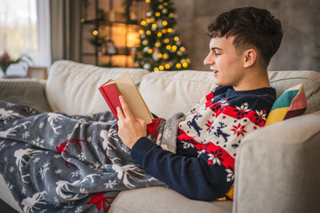 Young man lying down on sofa cover by festive blanket and read book