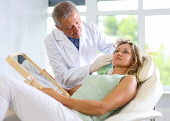Obraz premium Female patient of cosmetology office sitting in chair with mirror in hands and discussing with mature male doctor need for rejuvenation procedure laser skin resurfacing forehead
