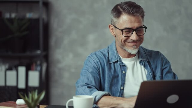 Portrait of casual good looking man smiling. Businessman sitting at desk in office drinking cup of coffee working on laptop. Successful, satisfied middle aged male entrepreneur at work.