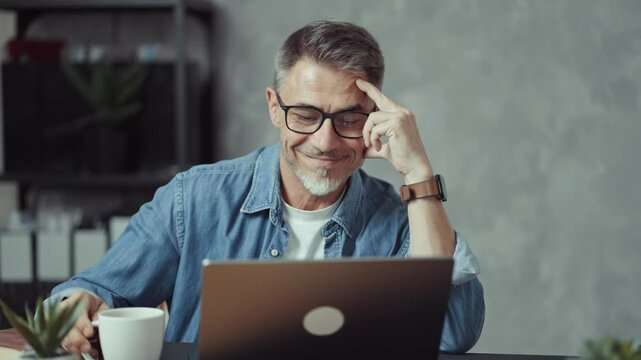 Portrait of mature man with grey hair and beard smiling. Confident businessman working on laptop from home office. Male entrepreneur sitting at desk in bright room wearing casual denim shirt.
