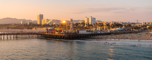 Santa Monica Pier extends into the Pacific Ocean, featuring a Ferris wheel and amusement park. The skyline and mountains are visible under a golden sunset glow.