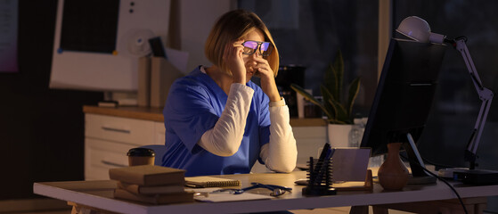 Tired female nurse with eyeglasses working evening shift at table in clinic
