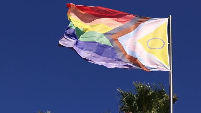 Intersex-inclusive redesign of the Progress Pride Flag. Rainbow LGBT flag against a blue sky. Flag fluttering in the wind. An abbreviation that stands for lesbian, gay, bisexual, transgender