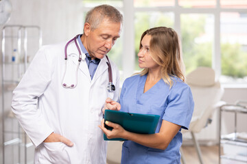 Middle-aged nurse and mature doctor discussing written diagnosis standing in doctor's cabinet
