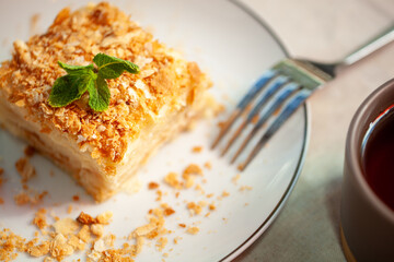 Partially eaten napoleon cake with a sprig of mint sits on a plate with crumbs and a fork, accompanied by a cup of tea