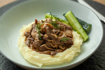Beef stroganoff served on a bed of mashed potatoes with a side of pickles and fresh dill. This dish is perfect for a hearty and satisfying meal