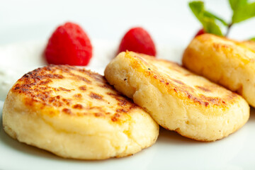 Cottage cheese pancakes with sour cream and raspberries are served on a white plate. The pancakes are decorated with mint