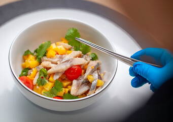 Chef wearing blue gloves is using tongs to carefully place a garnish on a gourmet salad made with fish, mango, tomatoes, and fresh herbs