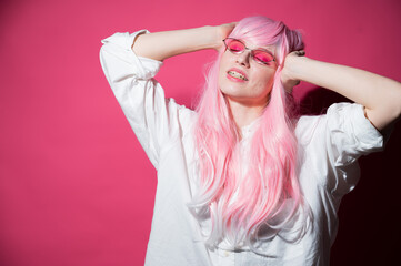 Naklejka premium Close-up portrait of a young woman with braces in a pink wig and sunglasses on a pink background. Copy space. 