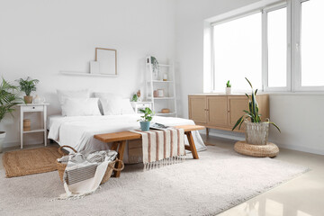 Stylish interior of light bedroom with wooden chest of drawers, bench, houseplants and bed