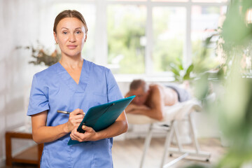 Masseuse writing at folder standing at massage room in front of male patient lying at massage table